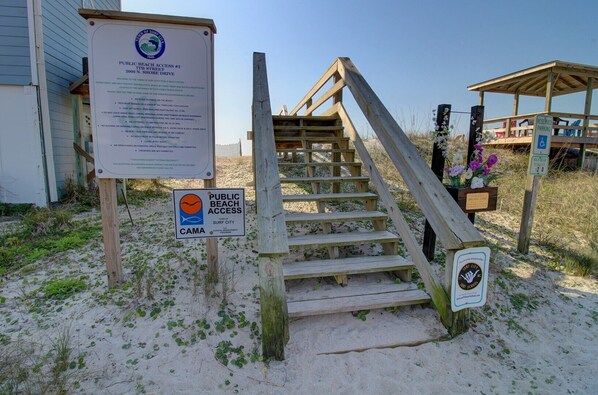 A wooden staircase leading to a beach access point with informational signs, a wheelchair-accessible ramp, and a small structure with flowers.