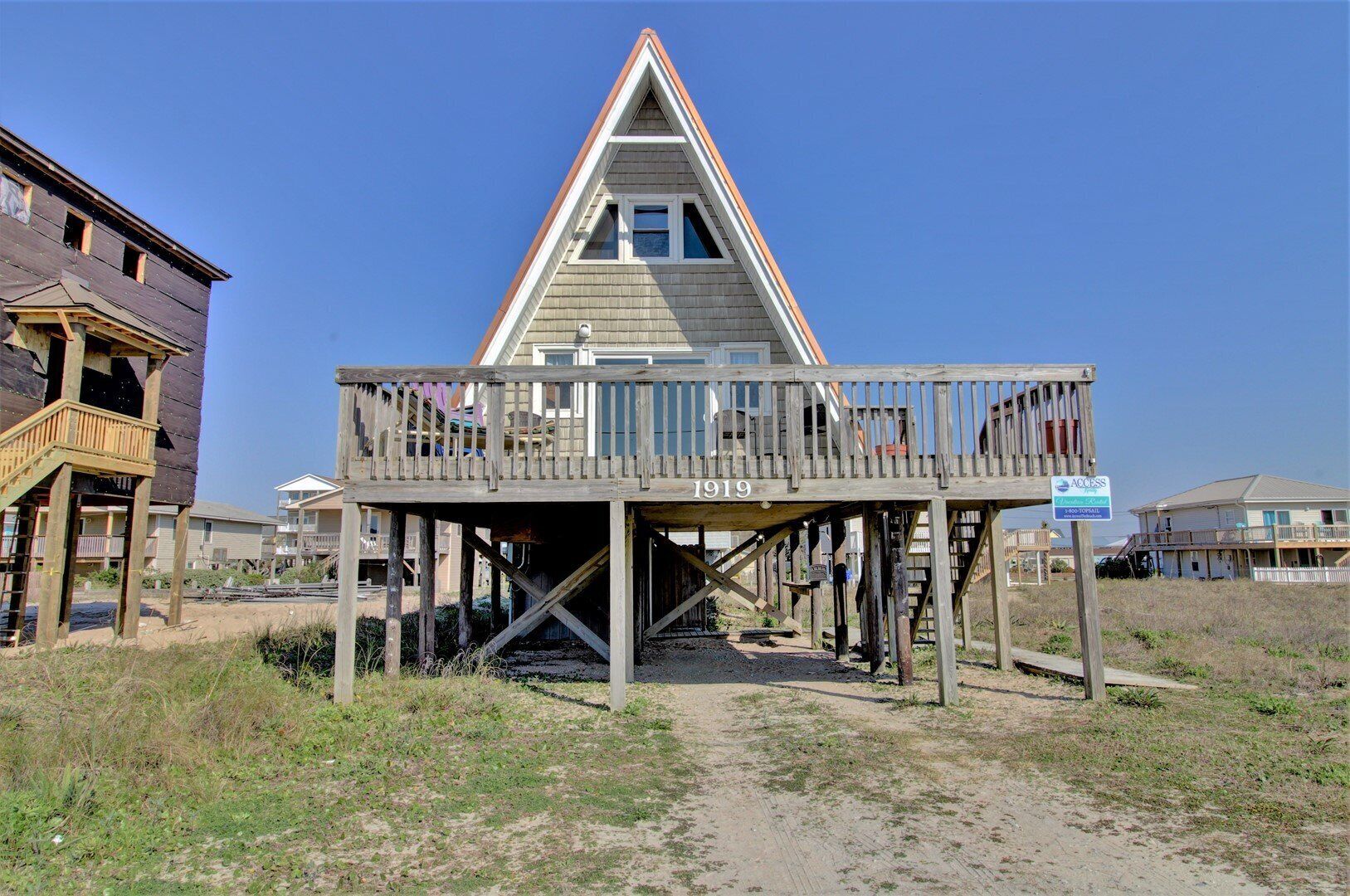 Une maison de plage en bois dotée d’un balcon, avec le numéro « 1919 » affiché sur la façade et un panneau indiquant « Accès à la partie supérieure de la plage ».