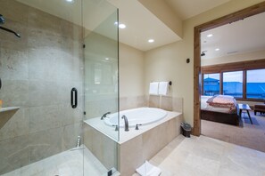 Een moderne badkamer met een douchecabine, een ruim bad en uitzicht op een slaapkamer met een bed en een tafel.