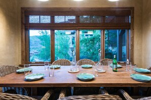 Een eettafel gedekt met borden, glazen en een fles, omringd door rieten stoelen, met uitzicht op groen door grote ramen.