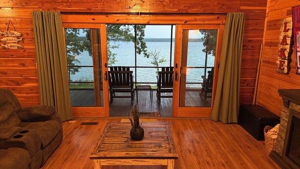 A cozy cabin interior with a wooden floor, a sofa, a coffee table with a plant, and a view of a lake through large glass doors.