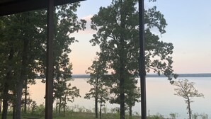 A view through a window with a metal frame, overlooking a calm lake with trees in the foreground and a distant shoreline.