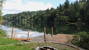 A lakeside deck with a fire pit, wooden dock, and a clear view of the lake surrounded by trees.