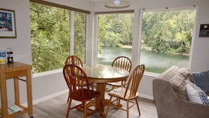 A dining area with a round table and four chairs, large windows with a view of a lake and trees, a wooden side table with a bottle, and a couch with pillows.