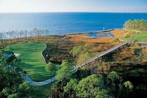 En golfbana med ett vattenhinder, en bro och utsikt över havet.