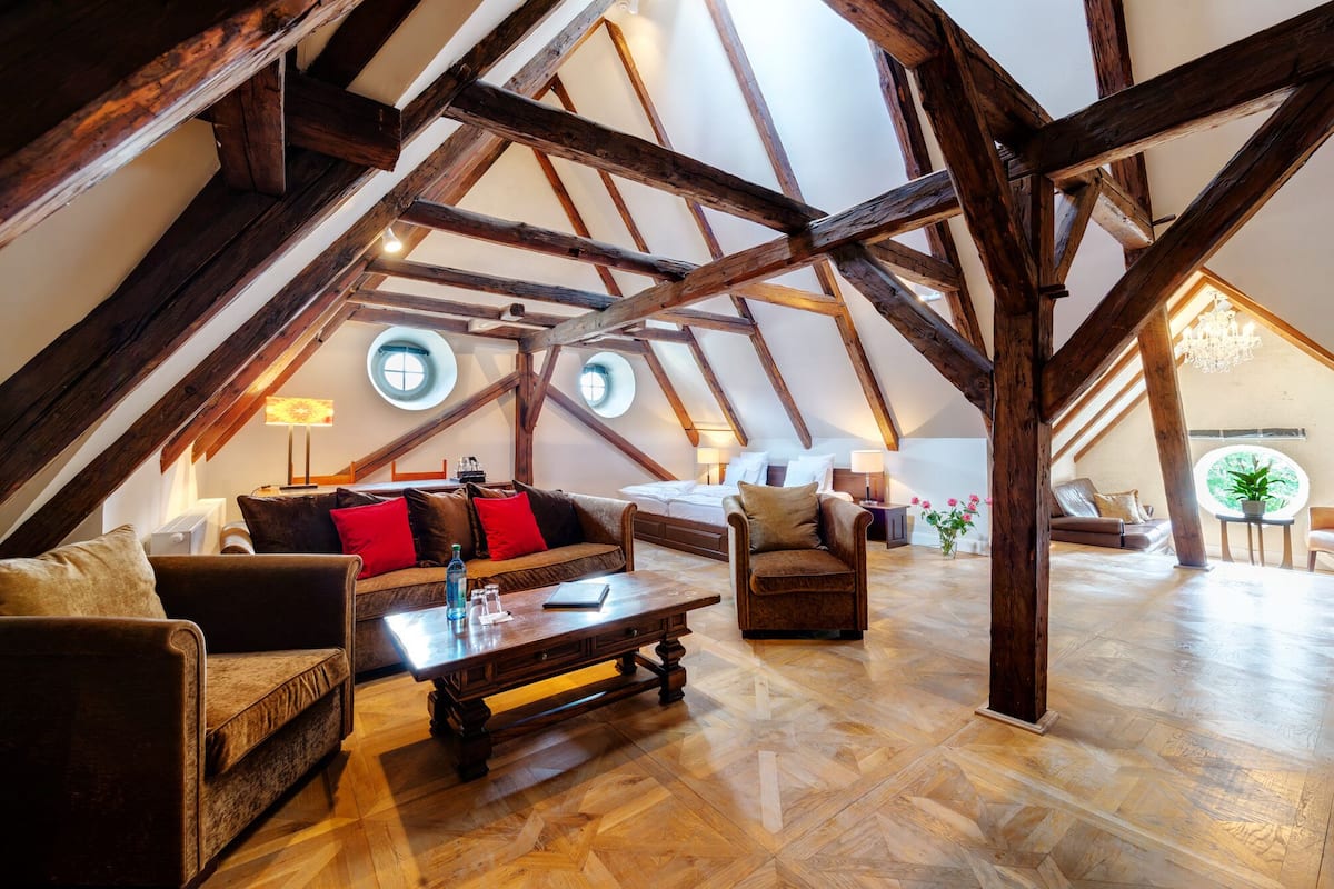 Een ruime loft met zichtbare houten balken, een groot rond raam en een gezellige zithoek met een bank en fauteuils.