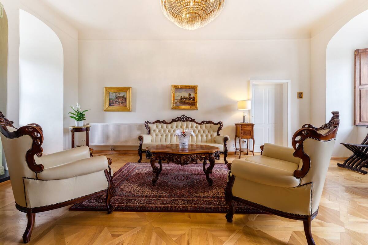 Een ruime woonkamer met een grote bank, twee fauteuils, een salontafel en een kroonluchter. Twee ingelijste schilderijen hangen aan de muur en op een bijzettafeltje staat een potplant.