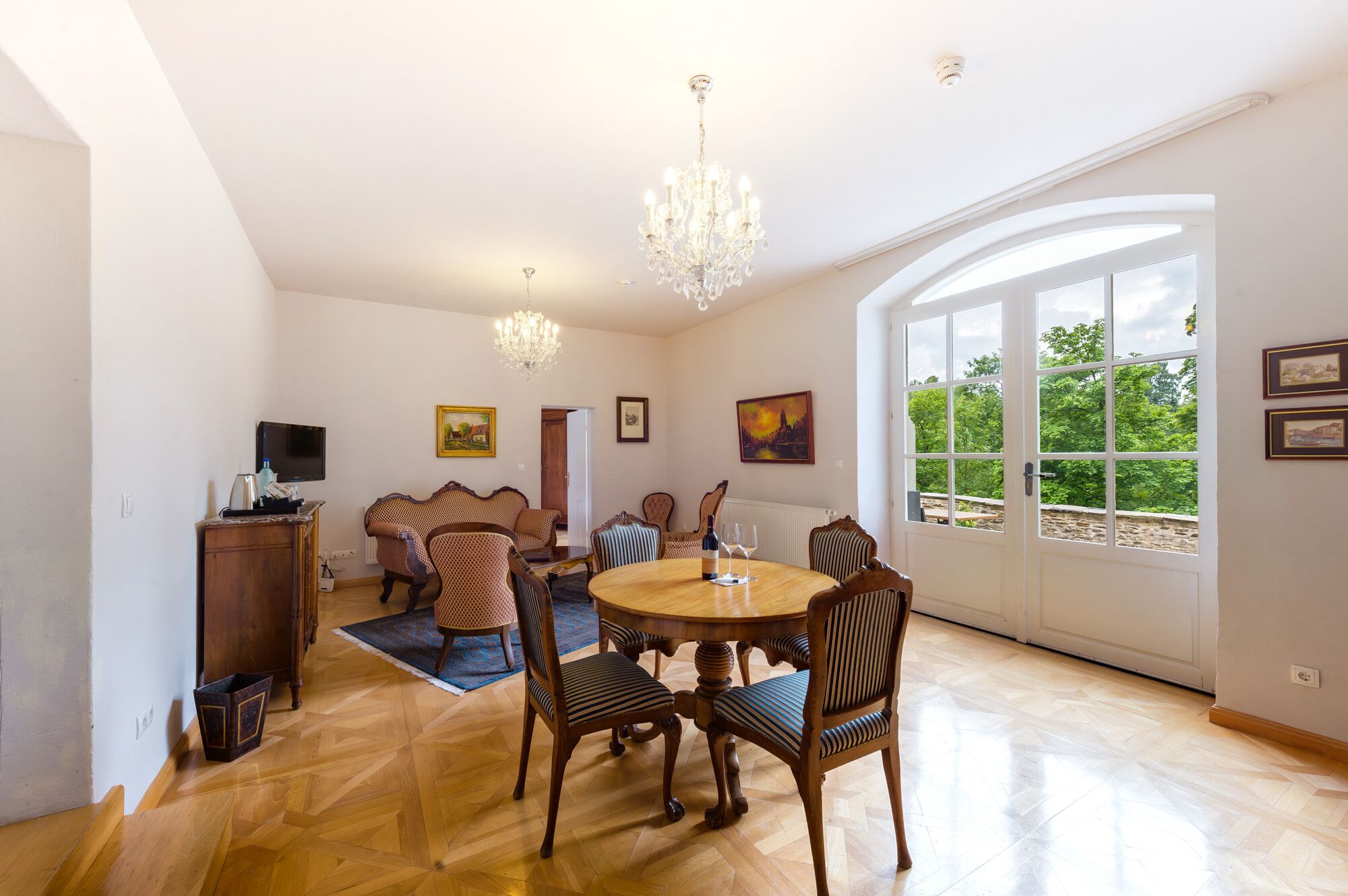 A dining room with a round wooden table, six chairs, a chandelier, and a large window with a view of greenery.