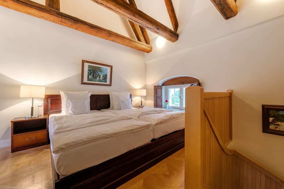 Een slaapkamer met een houten plafond, een bed met witte lakens, een nachtkastje met een lamp en een raam met uitzicht op groen.