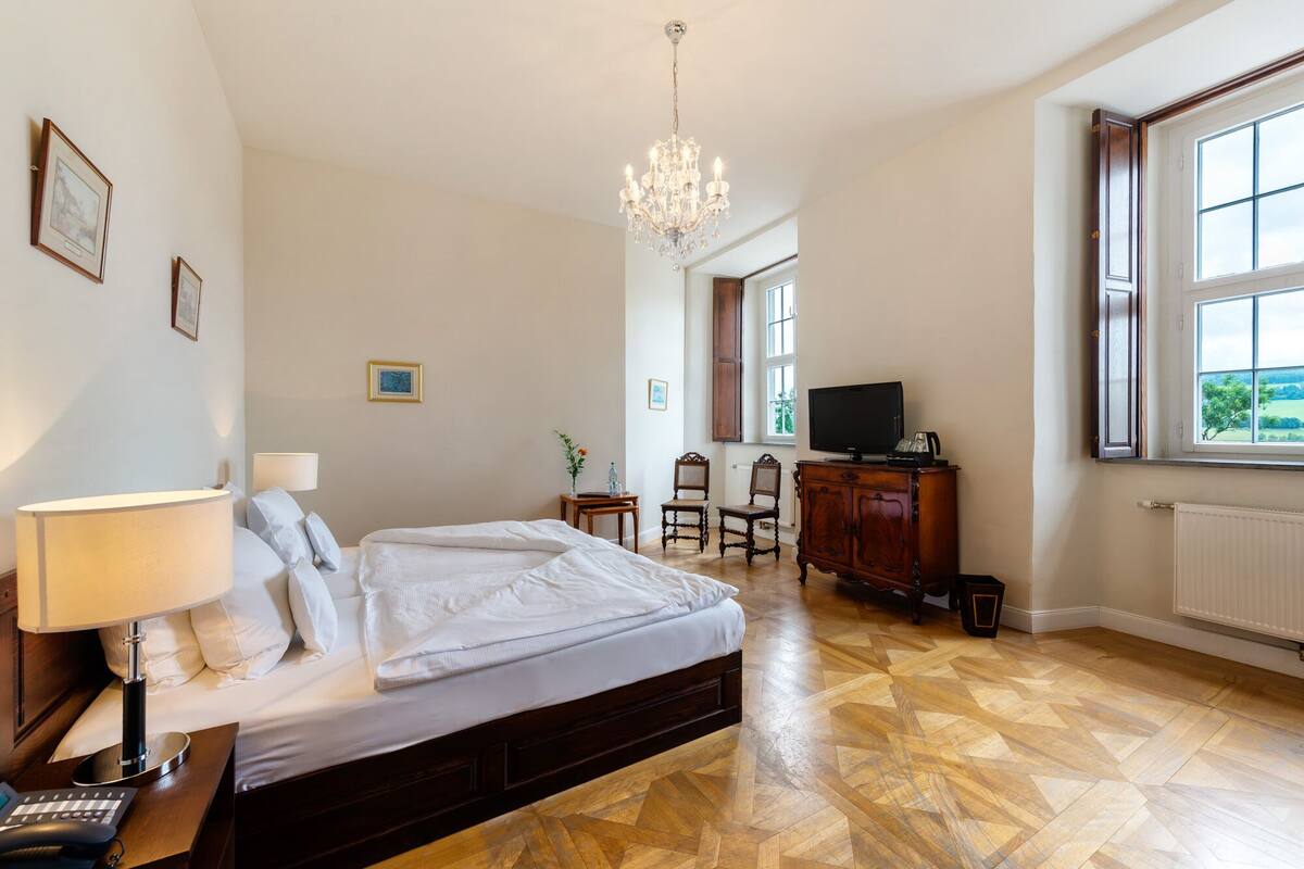 Een slaapkamer met een bed, een bureau, een stoel, een televisie en een raam met uitzicht op groen.