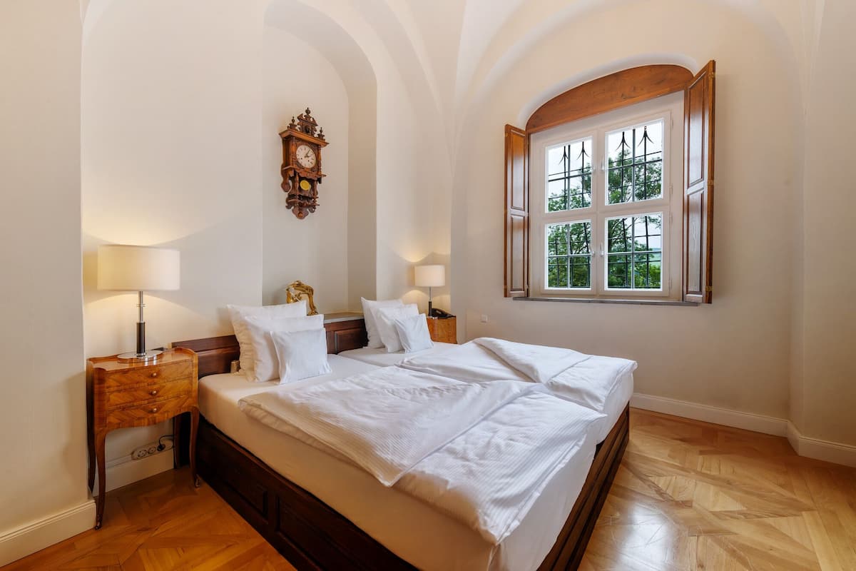 Een slaapkamer met een groot bed, twee nachtkastjes met lampen, een wandklok en een raam met uitzicht op bomen.