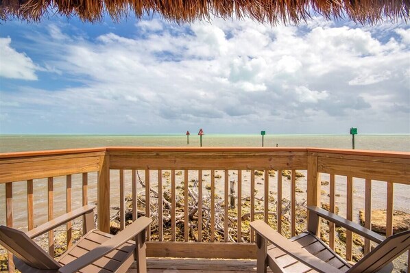 A wooden deck with two chairs facing a beach, with a thatched roof overhead.