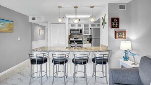 Una cocina moderna con armarios blancos, una isla central con taburetes de bar, iluminación colgante y decoración mural.