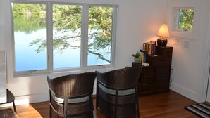Une pièce dotée d’une grande fenêtre donnant sur un lac, de fauteuils en osier, d’un meuble en bois rempli de livres et d’une table.