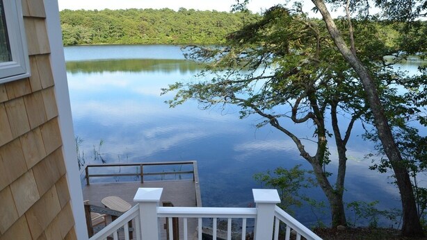 Une terrasse en bois dotée d’une balustrade, qui offre une vue sur un lac paisible, le tout entouré d’arbres.