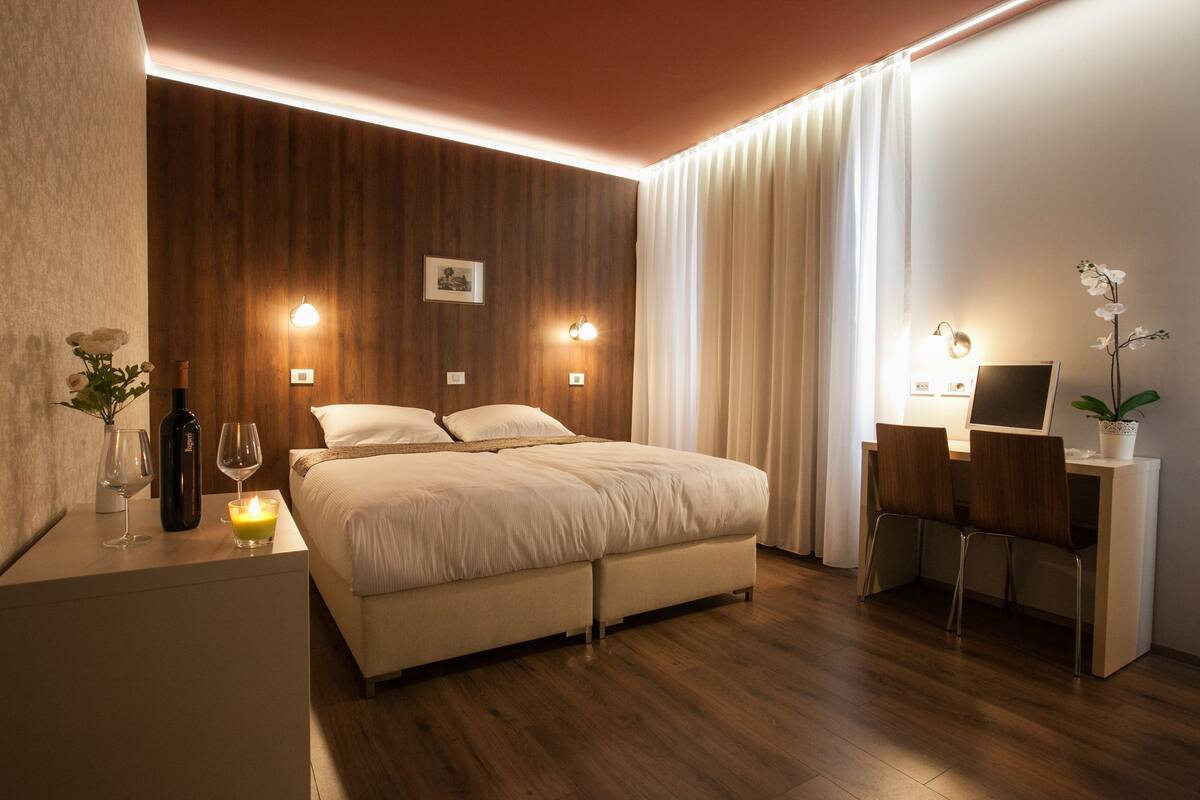 A modern bedroom with a wooden headboard, a bed with white bedding, a desk with a laptop, and a potted orchid.
