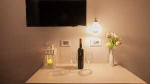 Ein Flachbildfernseher an der Wand, eine beleuchtete Tischlampe, eine Weinflasche, zwei Weingläser, ein Kerzen Laterne und eine Vase mit Blumen.