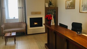 A living room with a fireplace, a striped sofa, a wooden coffee table, a reception desk, and a potted plant. - Hotel Center (Postojna)