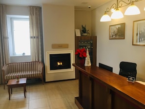 A living room with a fireplace, a striped sofa, a wooden coffee table, a reception desk, and a potted plant. - Hotel Center (Postojna)