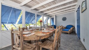 Une table de salle à manger en bois, mise avec des assiettes, des verres et une bougie. Les fenêtres sont ornées de rideaux bleus, et on y trouve une chaise en osier et un canapé bleu.