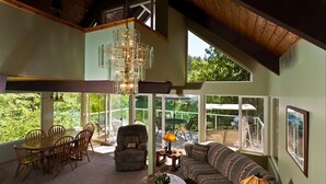 A spacious living area with a high ceiling, a chandelier, a sofa, a dining table with chairs, and large windows offering a view of greenery outside. - Emerald Lake Chalet - SUMMERTIME! (Bellingham)