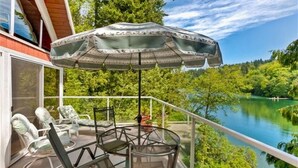 A balcony with a table and chairs, an umbrella, and a view of a lake surrounded by greenery. - Emerald Lake Chalet - SUMMERTIME! (Bellingham)
