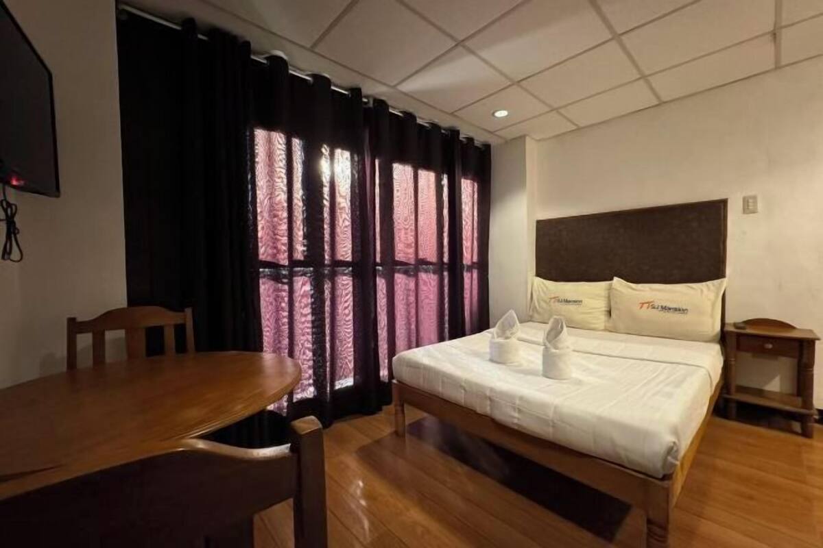 A hotel room with a bed, a wooden table, a chair, and a window with patterned curtains.