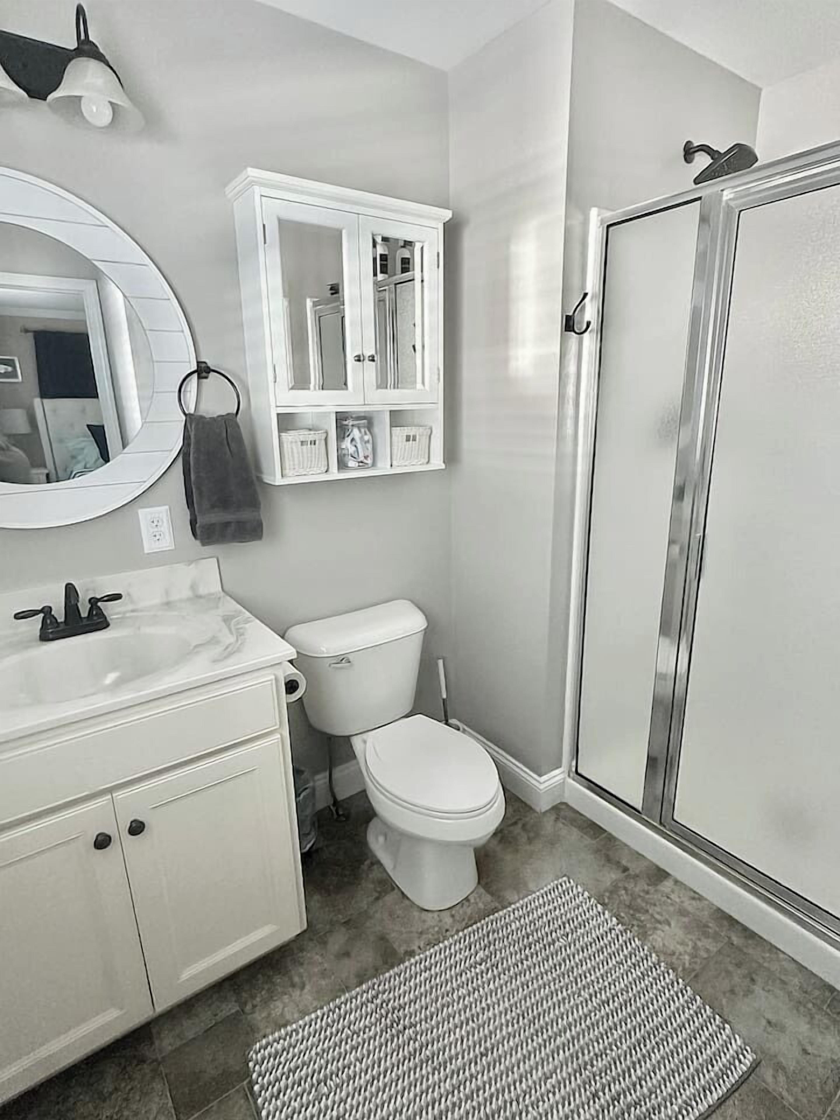 Een badkamer met een wit wastafel, toilet en douche. Boven het wastafel hangt een spiegel en er is een kastje met toiletartikelen.