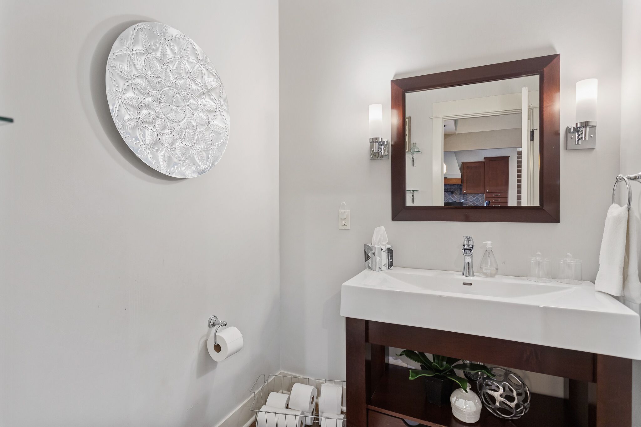 Un baño con un lavamanos blanco, un espejo grande, un dispensador de papel higiénico y un adorno de pared.