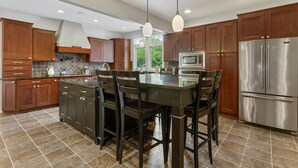 Une cuisine moderne avec des meubles en bois foncé, un grand îlot central doté de tabourets, des appareils électroménagers en acier inoxydable et une grande fenêtre qui laisse entrer la lumière naturelle.