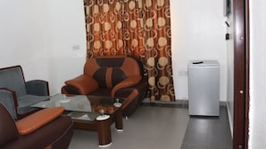 A living room with a sofa, armchair, and glass coffee table. There is a small refrigerator and a door to the right. -  Crystalem Luxury Hotel (Uyo)