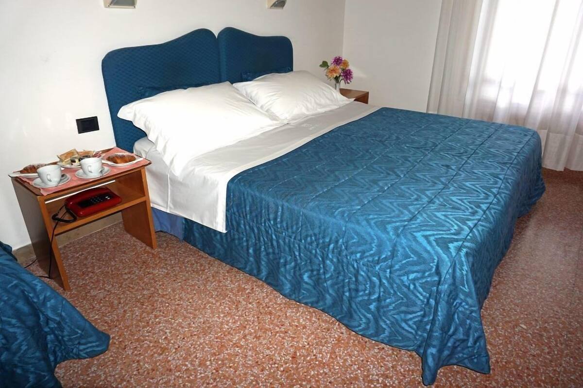 A hotel room with a bed, a nightstand with a tray of breakfast items, and a small table with a vase of flowers.