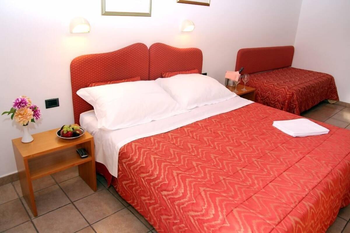 A hotel room with a large bed, a smaller bed, a nightstand with a fruit bowl, and a vase of flowers.