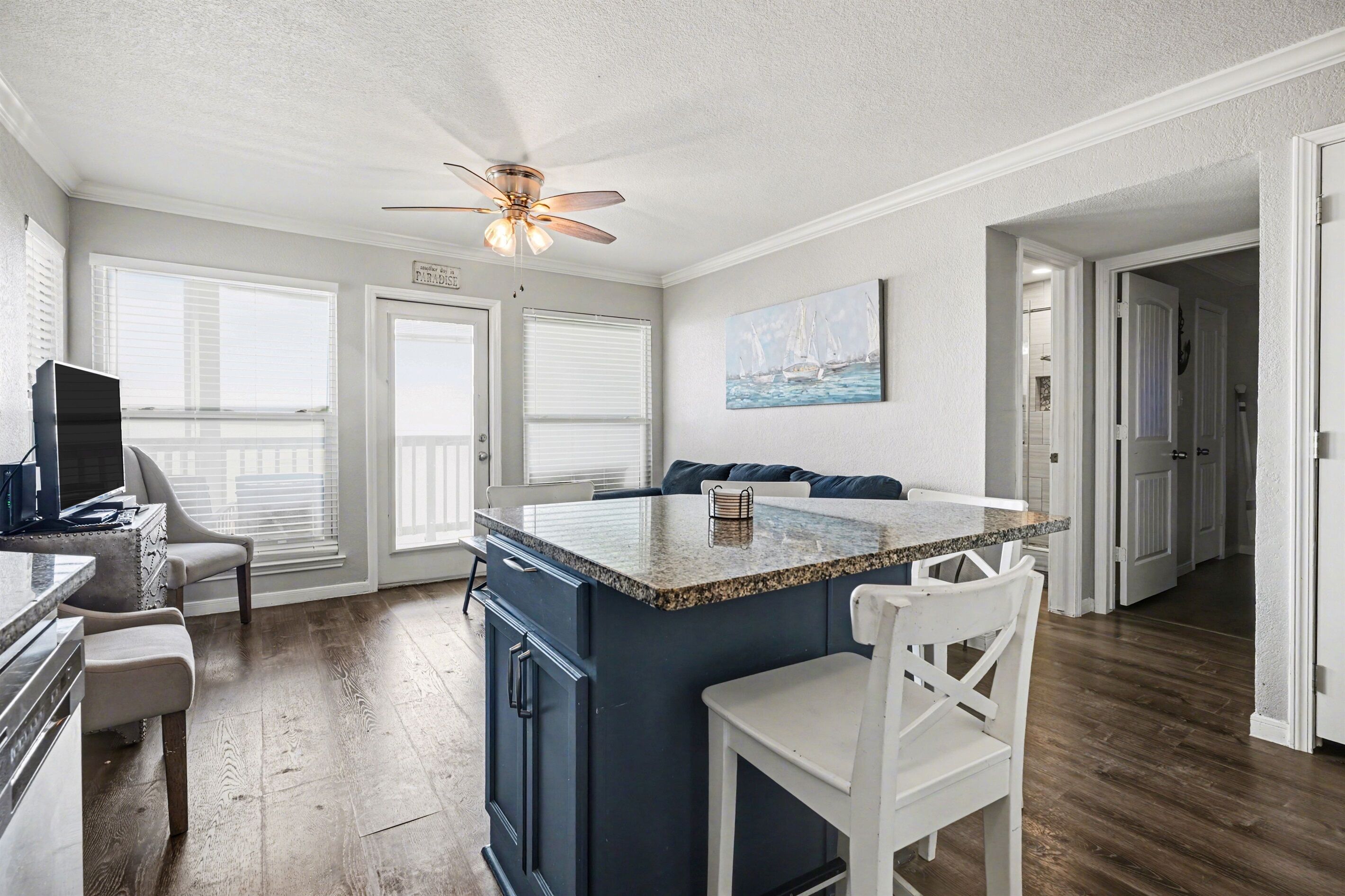 A kitchen with a central island, white chairs, a ceiling fan, and a painting on the wall.