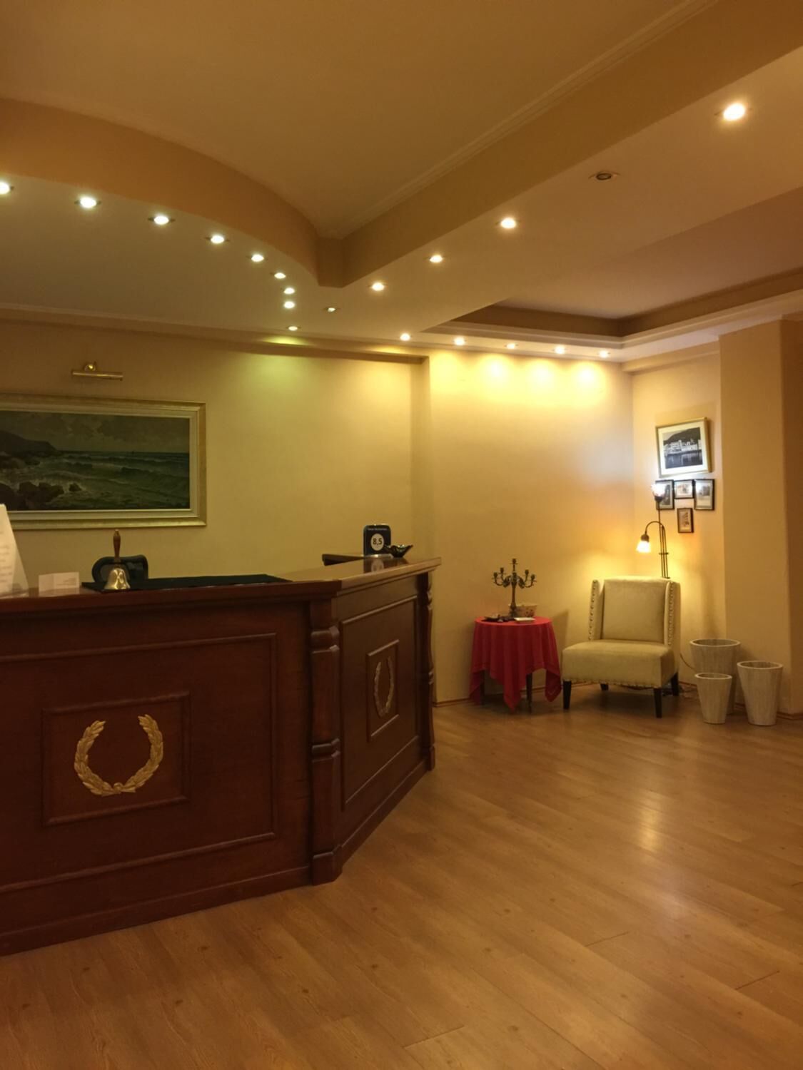 A room with a wooden reception desk, a painting on the wall, a small table with a red tablecloth, a chair, and a floor lamp.