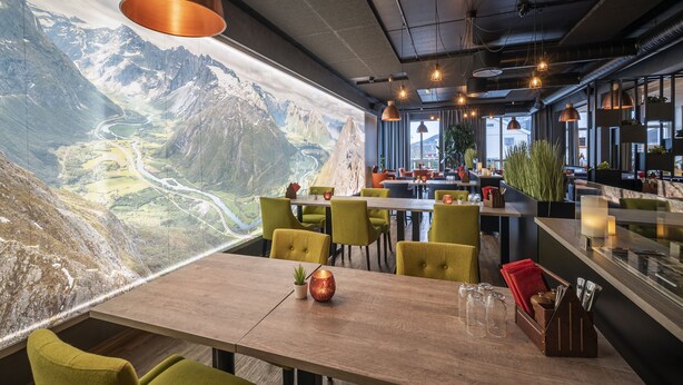 Un restaurante con un amplio ventanal que ofrece vistas a la montaña, mesas de madera, sillas verdes e iluminación colgante.