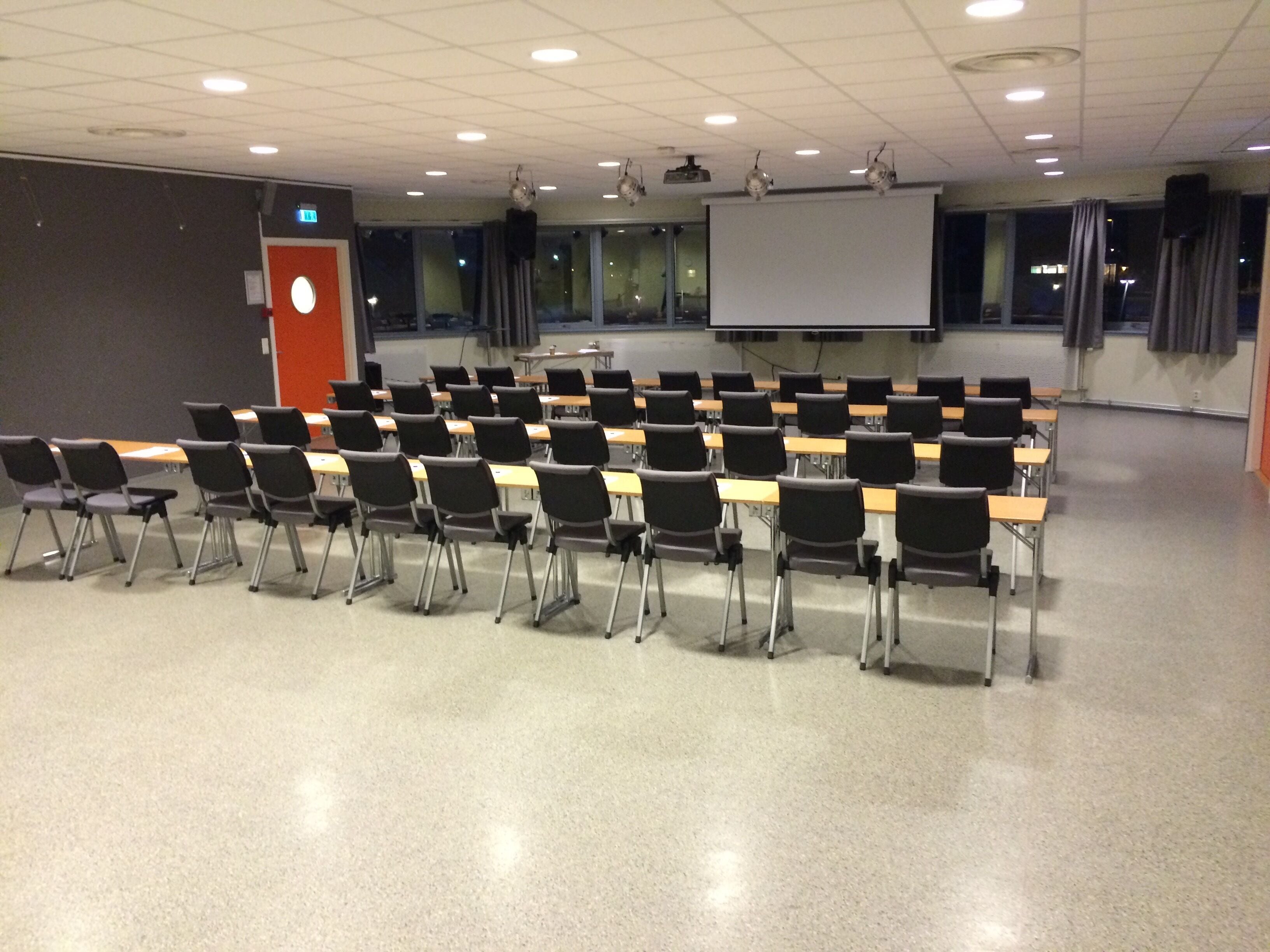 Een klaslokaal met rijen bureaus en stoelen, een scherm voor een projector en een whiteboard.