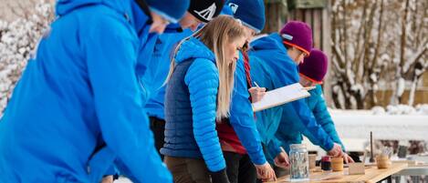 A group of people in blue jackets and hats gathered around a table with various items, possibly for a workshop or activity, outdoors in a snowy setting.