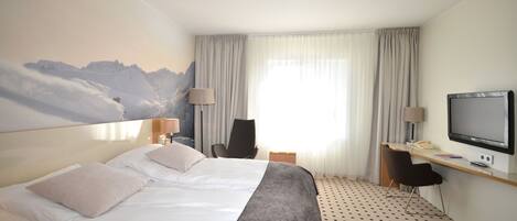 Quarto de hotel com uma cama grande, uma escrivaninha com televisão, uma cadeira e vista para as montanhas pela janela.