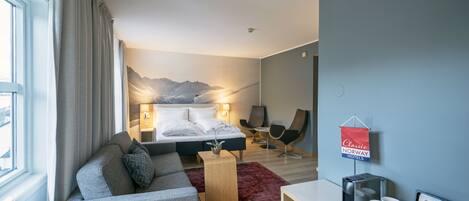A modern hotel room with a bed, a sofa, a desk, and a chair. There is a window with curtains, a wall-mounted sign, and a view of mountains.