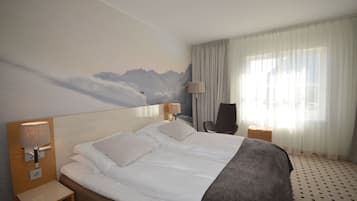 A hotel room with a large bed, a nightstand with a lamp, a chair, and a window with sheer curtains.
