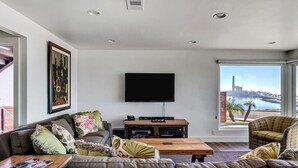 Ein modernes Wohnzimmer mit einem großen Flachbildfernseher, einer Sofagarnitur, einem Couchtisch und Blick auf ein Gewässer durch große Fenster.