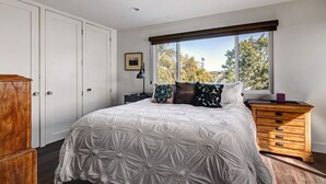Ein Schlafzimmer mit einem großen Bett, einem hölzernen Schminktisch, einem Nachttisch, einem Deckenventilator und einem Fenster mit Blick auf Bäume.