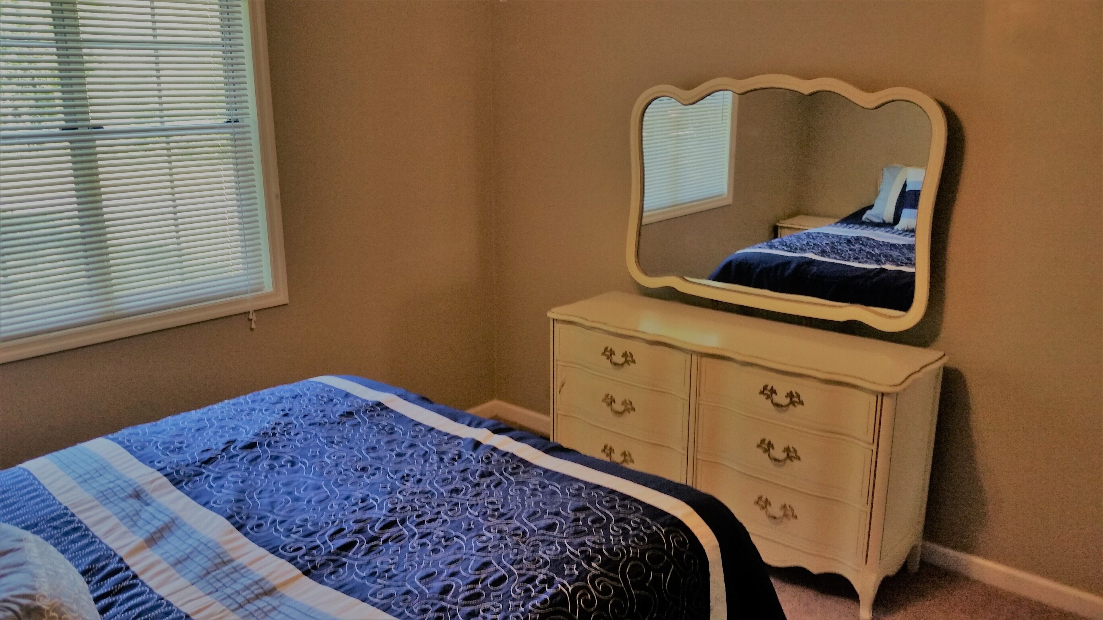 A bedroom with a bed, a dresser, a mirror, and a window with blinds.