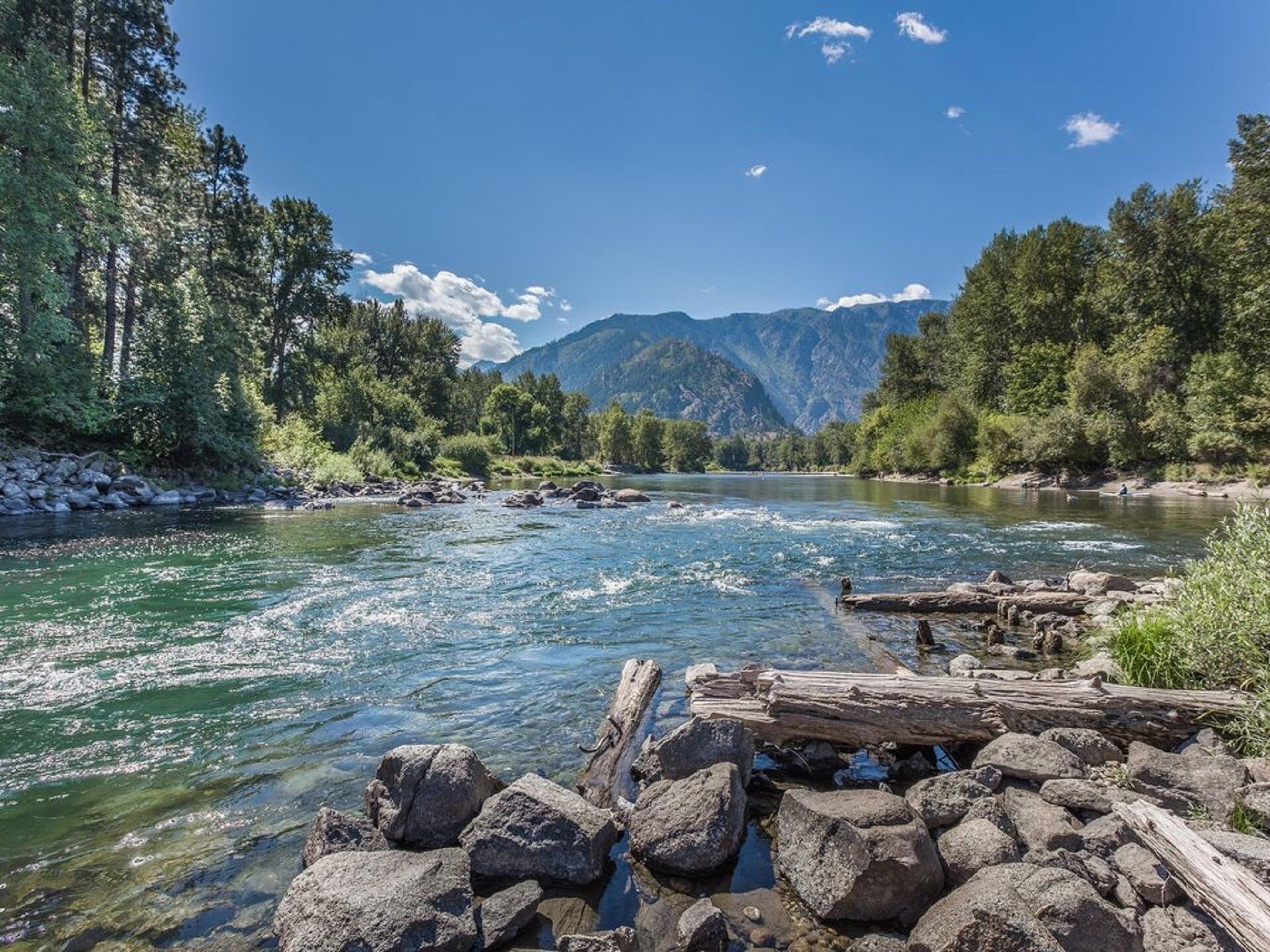 Une rivière aux eaux claires, bordée de rochers et d’arbres, avec des montagnes en arrière-plan.
