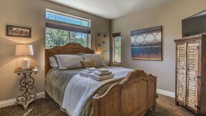 Un dormitorio con cama de madera, una mesita de noche con lámpara, ventana con vista a árboles y un cuadro en la pared.