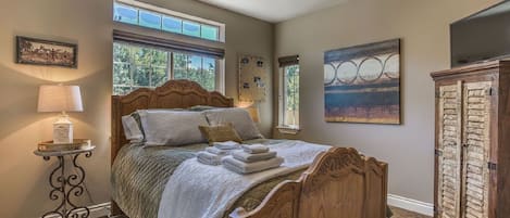 A bedroom with a wooden bed, a nightstand with a lamp, a window with a view of trees, and a painting on the wall.