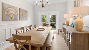 A dining room with a wooden table, chairs, a chandelier, artwork, and a view through a door.