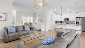 A modern living room with a grey sofa, a coffee table, and a kitchen area with white cabinets and stainless steel appliances.