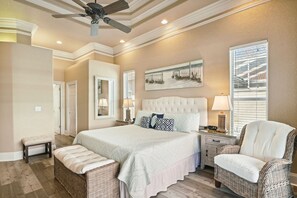 A bedroom with a large bed, two bedside tables, a chair, a bench, a ceiling fan, and a painting on the wall.